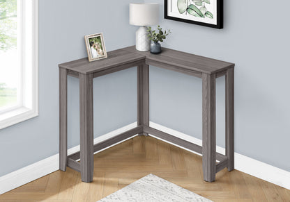 The Corner Accent Console Table For Entryway, Unique Design Contemporary & Modern, is a small gray L-shaped table with a lamp, framed photo, and plant on top, placed by a window on a wood herringbone floor against light gray walls and near a rug.