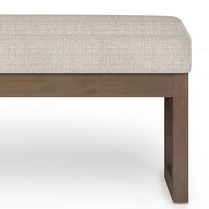 A close-up of the Milltown - Upholstered Ottoman Bench, featuring a light beige cushion and wooden square-leg base, set against a white background.