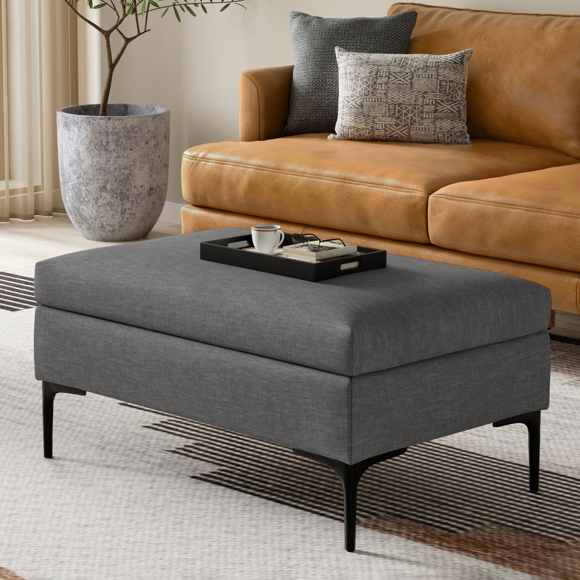 The Rebecca Multifunctional Coffee Table Storage Ottoman with Lift Up Lid, in gray upholstery and black legs, sits on a patterned rug before a tan leather sofa, topped with a tray holding coffee essentials. A large plant in a stone pot is behind.