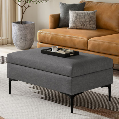 The Rebecca Multifunctional Coffee Table Storage Ottoman with Lift Up Lid, in gray upholstery and black legs, sits on a patterned rug before a tan leather sofa, topped with a tray holding coffee essentials. A large plant in a stone pot is behind.
