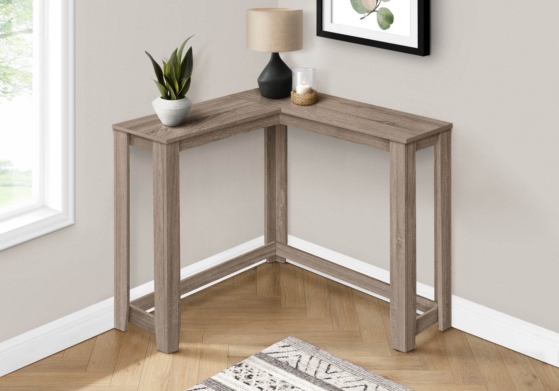 The Corner Accent Console Table For Entryway, Unique Design Contemporary & Modern, is an L-shaped wooden table styled with a plant, lamp, and candle in a light room featuring a framed picture on the wall and a rug over the wooden floor.