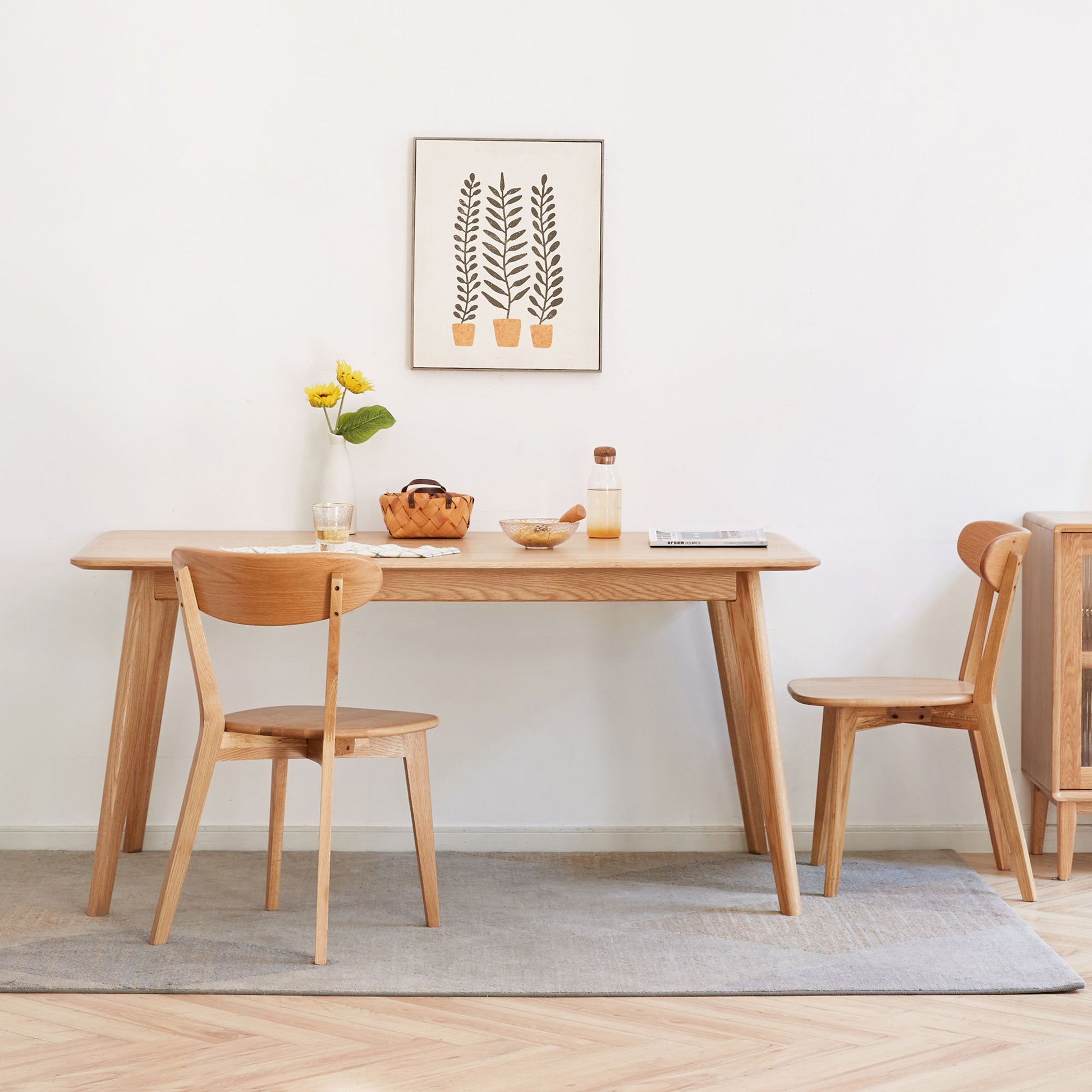 A minimalist dining area featuring a wooden table and two Simple Dining Chairs, with a small vase of yellow flowers, a fruit bowl, and framed plant art on a white wall, all atop a light grey rug.