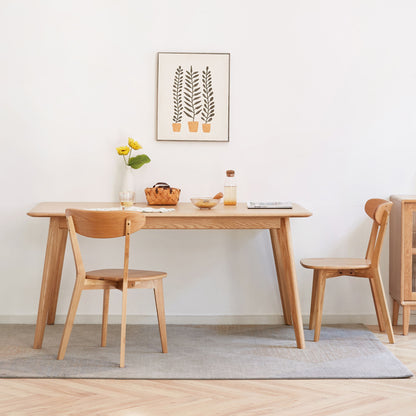 A minimalist dining area featuring a wooden table and two Simple Dining Chairs, with a small vase of yellow flowers, a fruit bowl, and framed plant art on a white wall, all atop a light grey rug.