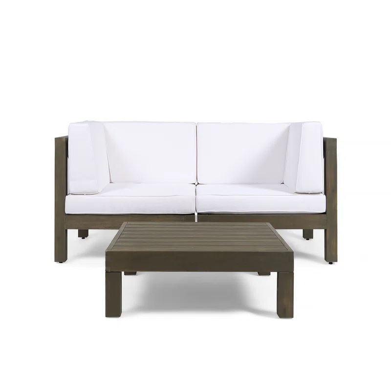 The Oana 3 Piece Love Seat Set features a modern outdoor sofa with white cushions and a dark wooden frame, paired with a matching low rectangular coffee table, all displayed against a plain white background.