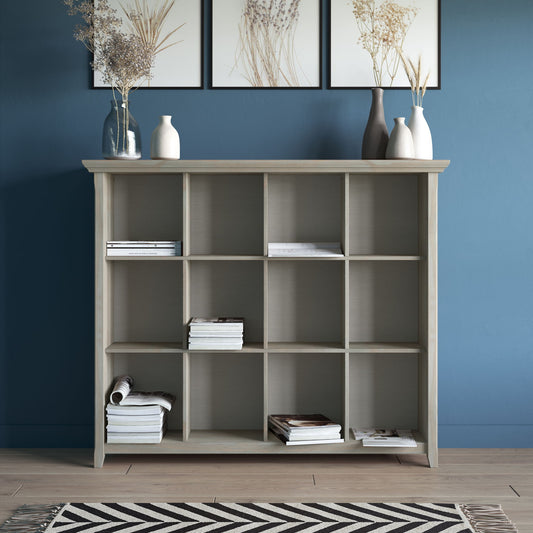 The Acadian - Handcrafted Cube Storage, a light wood bookshelf with twelve cubbies, stands against a blue wall, partially filled with books. Decorative vases with dried grasses sit on top and a geometric rug lies on the floor.