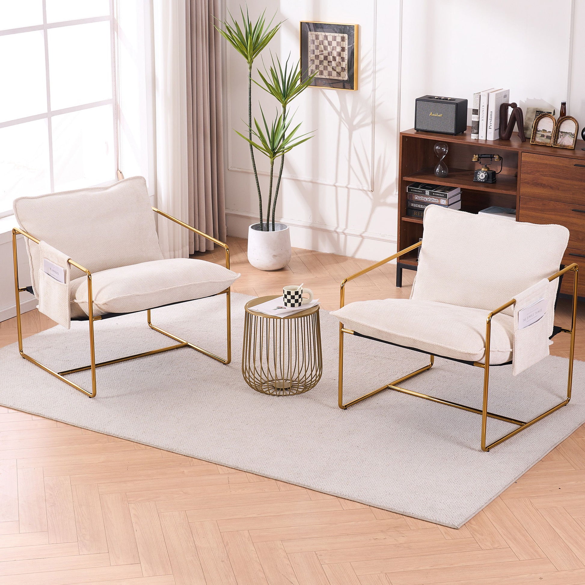Two Upholstered Hanging Armchairs with gold-plated metal frames, crushed foam cushions, and skin-friendly fabric sit on a light rug by a round table with a chessboard. A tall plant and bookshelf decor are near large curtained windows in the background.