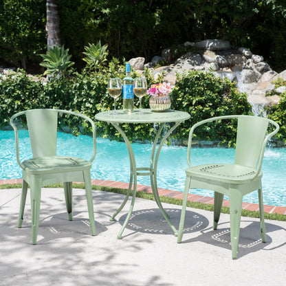 The Ambrosia 3 Piece Bistro Set with leaf motifs, featuring two light green metal chairs and a matching round table, is poolside holding wine glasses, a bottle, and pink flowers, with lush greenery and rocks behind.