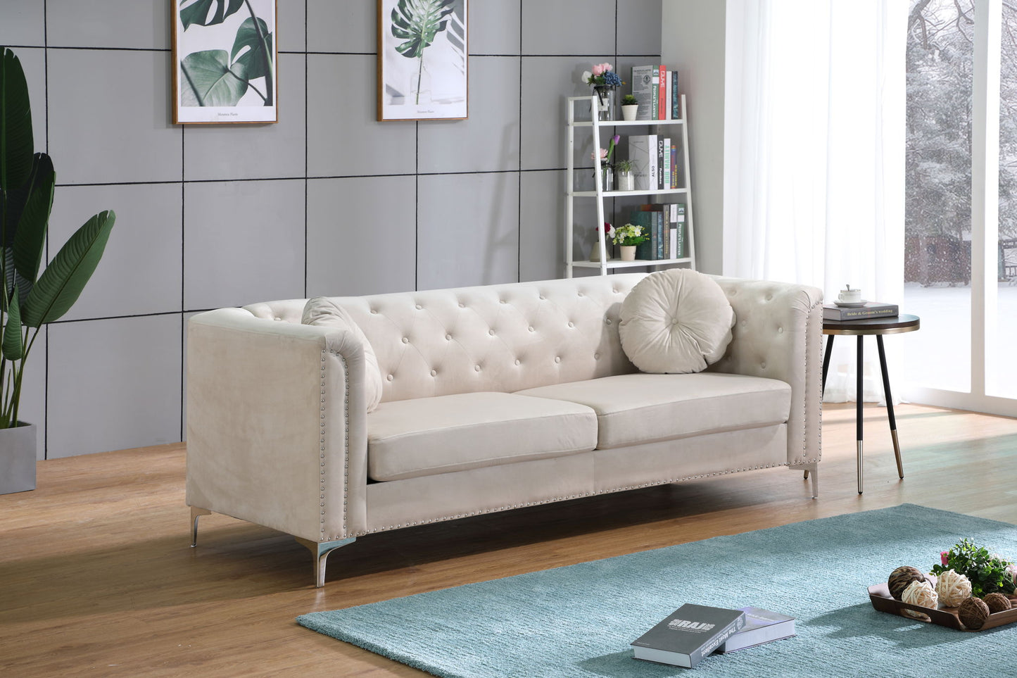 A modern living room features an Elegant Contemporary Sofa in light beige, a round pillow, blue rug, books, and a potted plant. The space is finished with framed botanical prints, a white bookshelf, and a large window with sheer curtains.