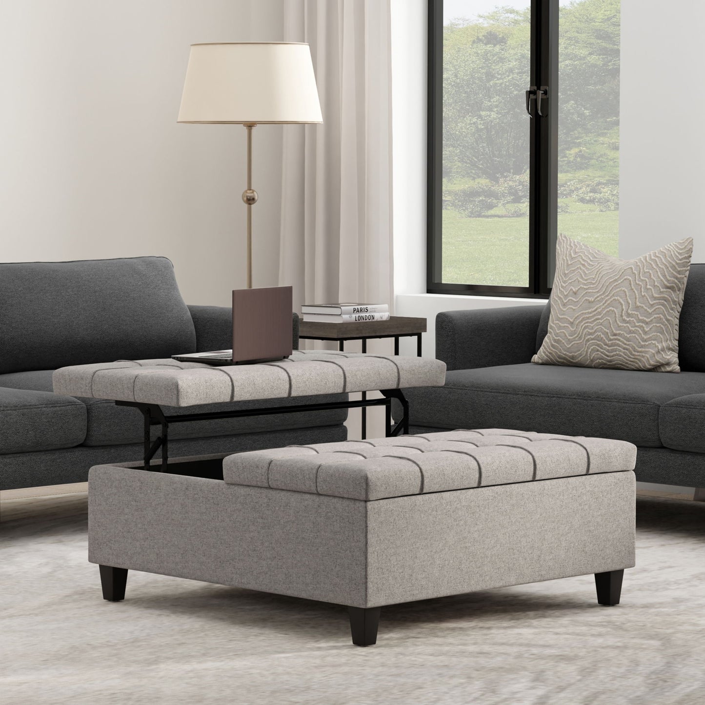 A modern living room with gray sofas, neutral decor, light curtains, and a patterned pillow features the Harrison Upholstered Coffee Table Storage Ottoman with a tufted gray lift-top tray and a small side table holding a book and lamp by the large window.