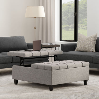 A modern living room with gray sofas, neutral decor, light curtains, and a patterned pillow features the Harrison Upholstered Coffee Table Storage Ottoman with a tufted gray lift-top tray and a small side table holding a book and lamp by the large window.
