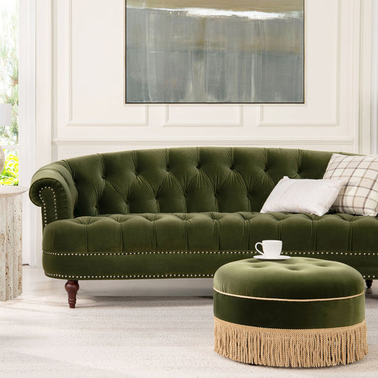 The La Rosa - Victorian Chesterfield Tufted Sofa in green velvet with studded trim is paired with a round fringed ottoman. Two pillows and a teacup rest on them, while a large abstract painting decorates the wall behind.