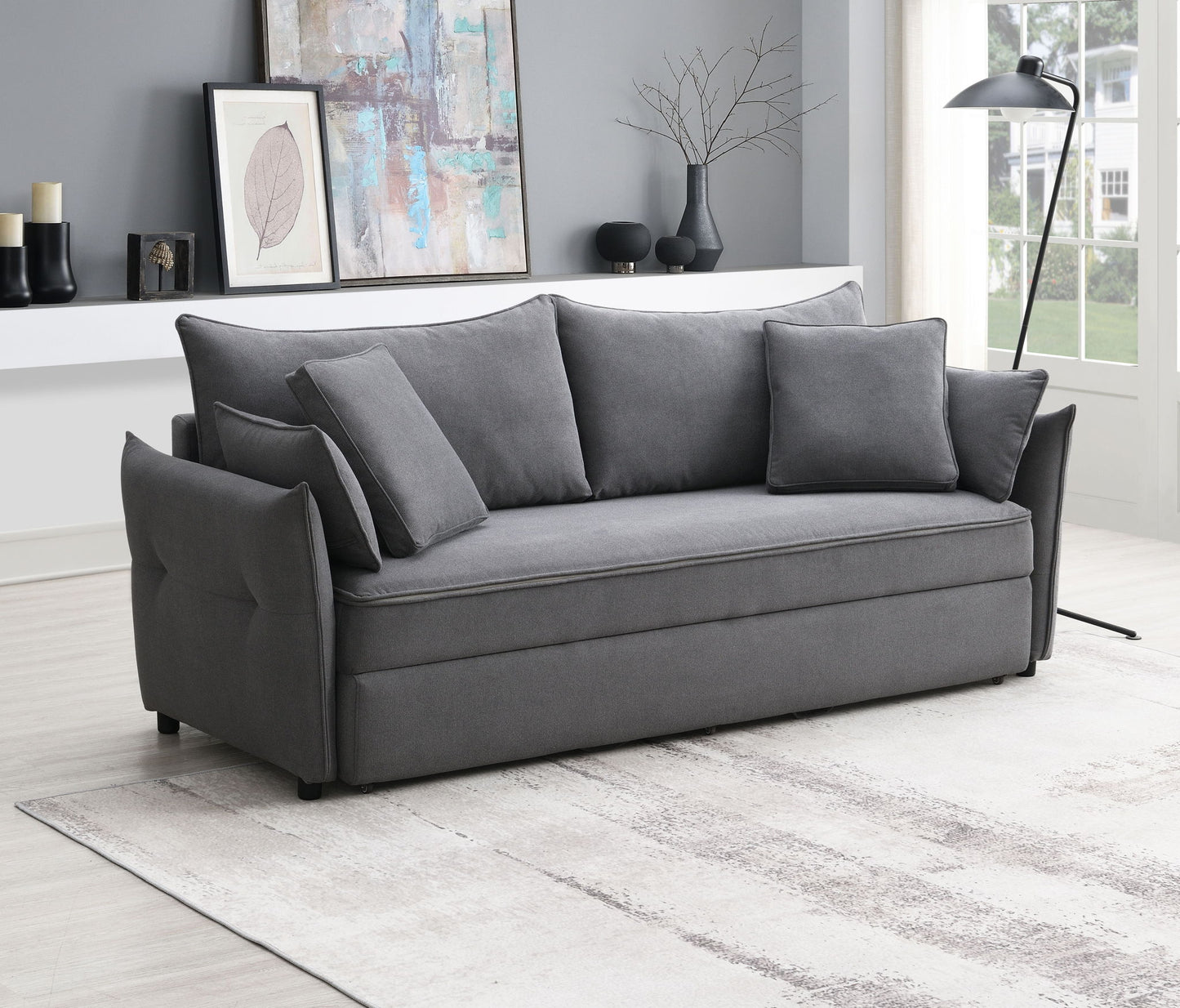 A modern living room with the Irina Pull Out Sleep sofa in gray (three back cushions, two side pillows), a light patterned rug, abstract wall art, a floor lamp, and a shelf with decor against gray walls.