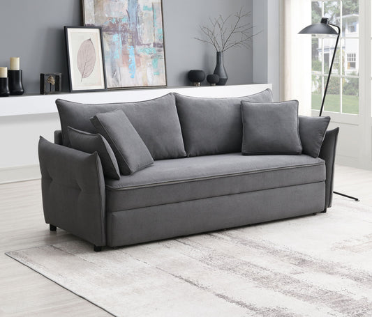 A modern living room with the Irina Pull Out Sleep sofa in gray (three back cushions, two side pillows), a light patterned rug, abstract wall art, a floor lamp, and a shelf with decor against gray walls.