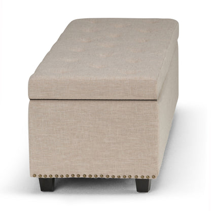 The Hamilton Upholstered Storage Ottoman features a rectangular beige design, button-tufted top, small brass nailhead trim at the base, and short dark wooden legs, shown from the front.