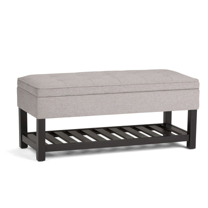 The Cosmopolitan Storage Ottoman Bench features a light gray cushioned top, black wooden legs, and an open slatted shelf underneath for added storage.