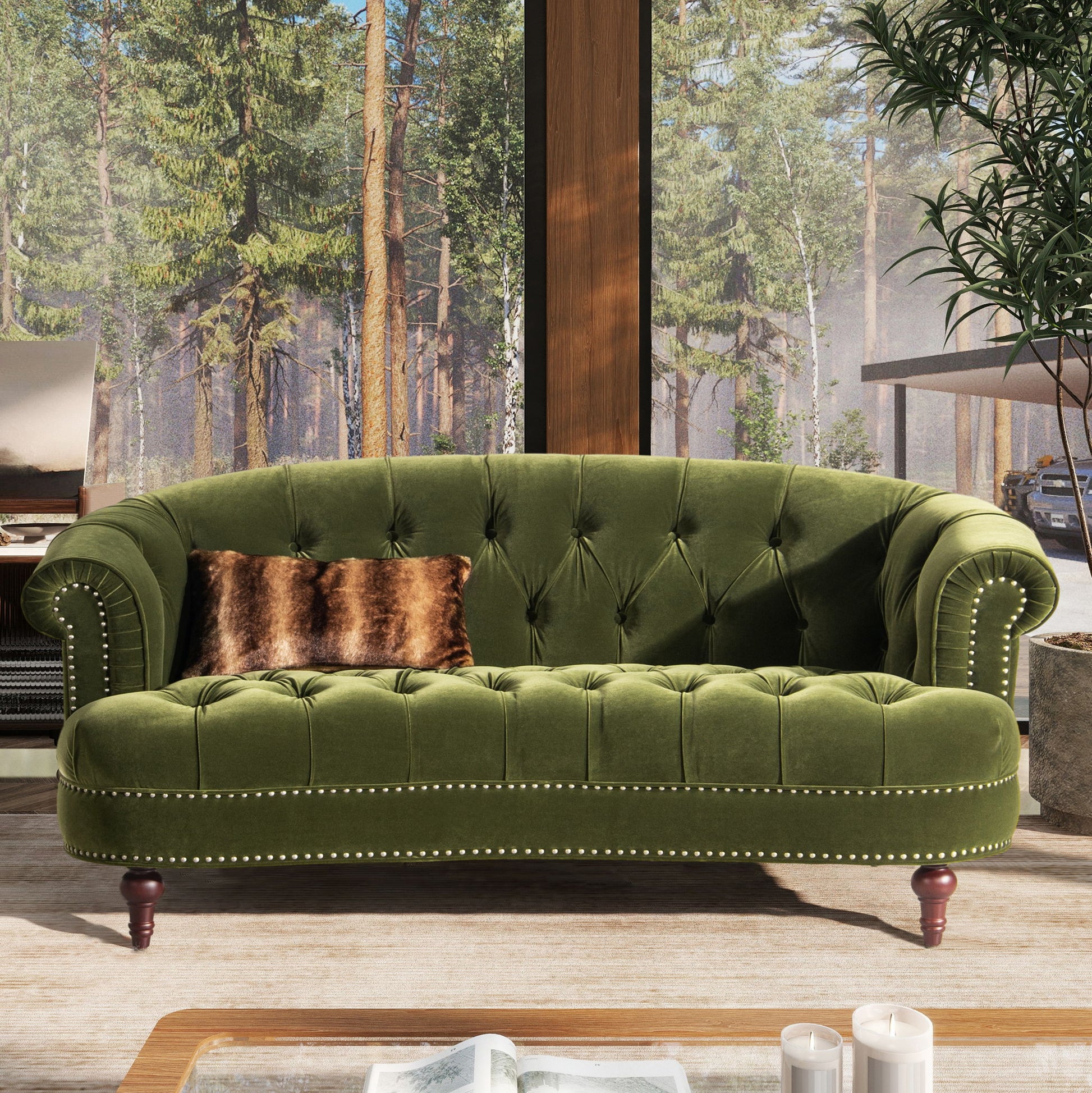 The La Rosa - Victorian Chesterfield Loveseat in green velvet with nailhead trim sits in a sunlit room, accented by a brown furry pillow. Large windows display forest views, bringing nature into the cozy space.