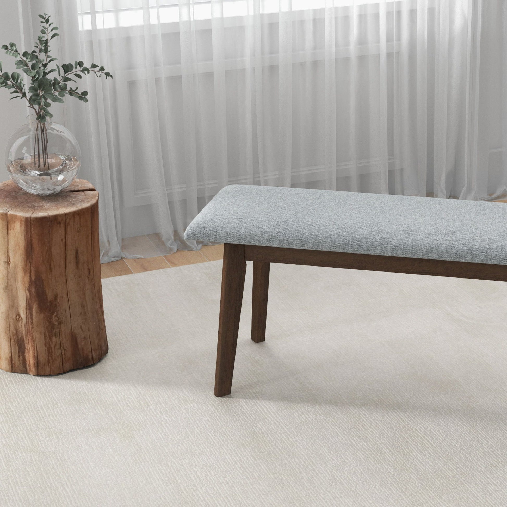 A minimalist room features the Carlos - Upholstered Bench with wooden legs, a wooden stump side table topped with a glass vase of greenery, and sheer white curtains over the window.