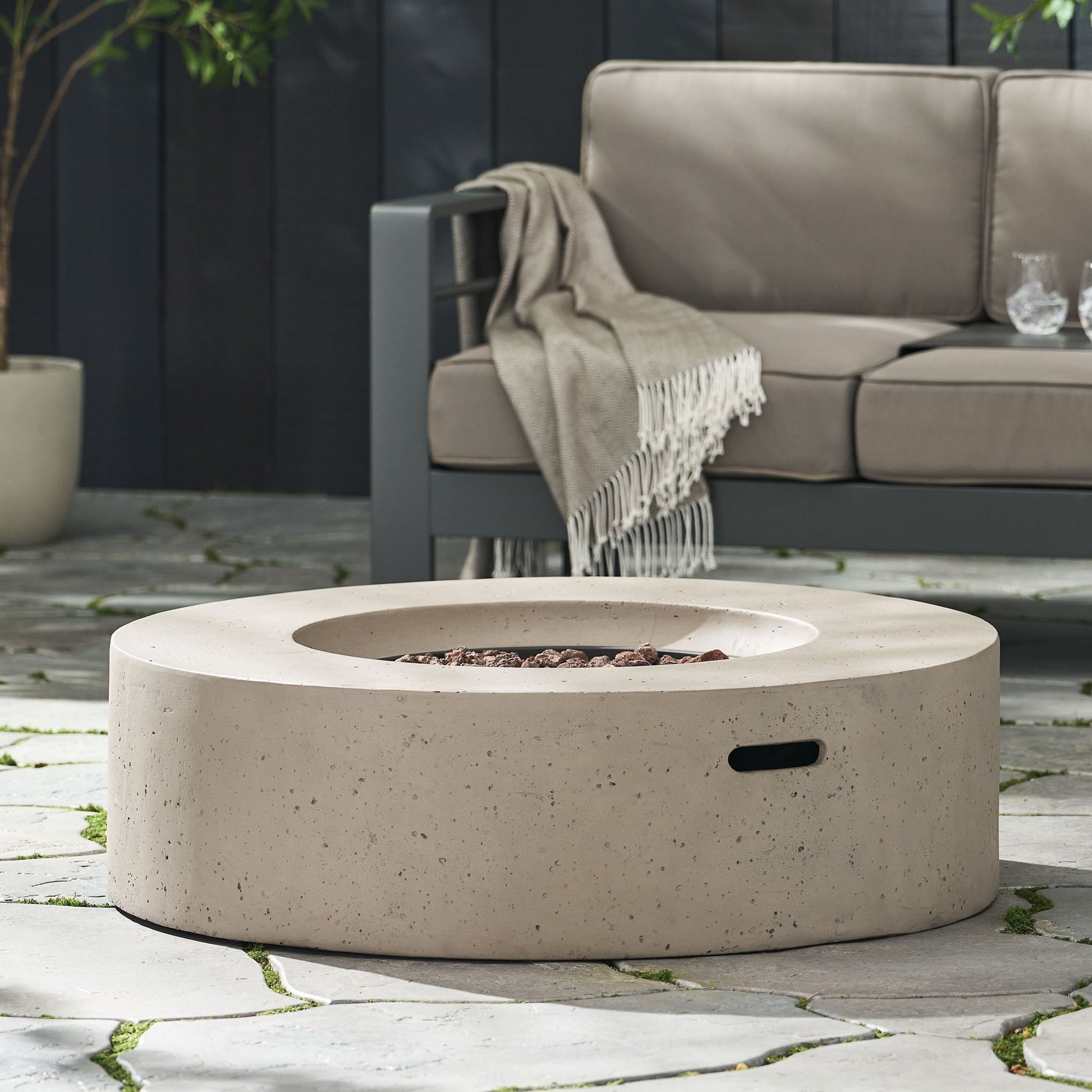 The Modern Lightweight Concrete Outdoor Circular Fire Pit 50,000 Btu sits on a stone patio beside an outdoor sofa with a beige throw and two glasses on the armrest.