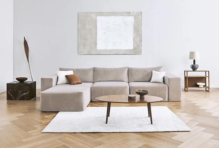 A modern living room with a beige sectional sofa adorned with white and rust-colored pillows. There's a wooden coffee table on a white rug, abstract wall art above the sofa, a dark side table with a sculpture on the left, and a wooden shelf with a lamp on the right.