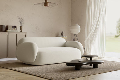 A cozy, modern living room features the Dori Sofa by Acanva on a textured rug. A dark, low coffee table is positioned in front. Natural light flows through sheer curtains, illuminating a cabinet with decor items and a curved floor lamp against the wall.