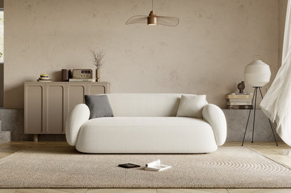 A cozy living room features the Dori Sofa by Acanva, complemented by two cushions. Behind it stands a wooden cabinet adorned with decor items and a vintage radio. A woven rug covers the floor, with a phone and a book resting on it. Overhead, a pendant light illuminates the space.