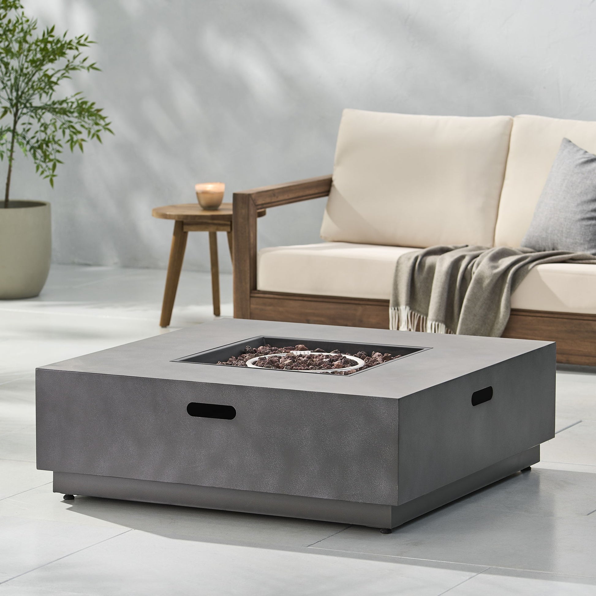 A modern outdoor patio with a Modern Square Fire Pit With 50,000 Btu, a light wood sofa with cream cushions, a throw blanket, a wooden side table holding a candle, and a potted plant in the background.