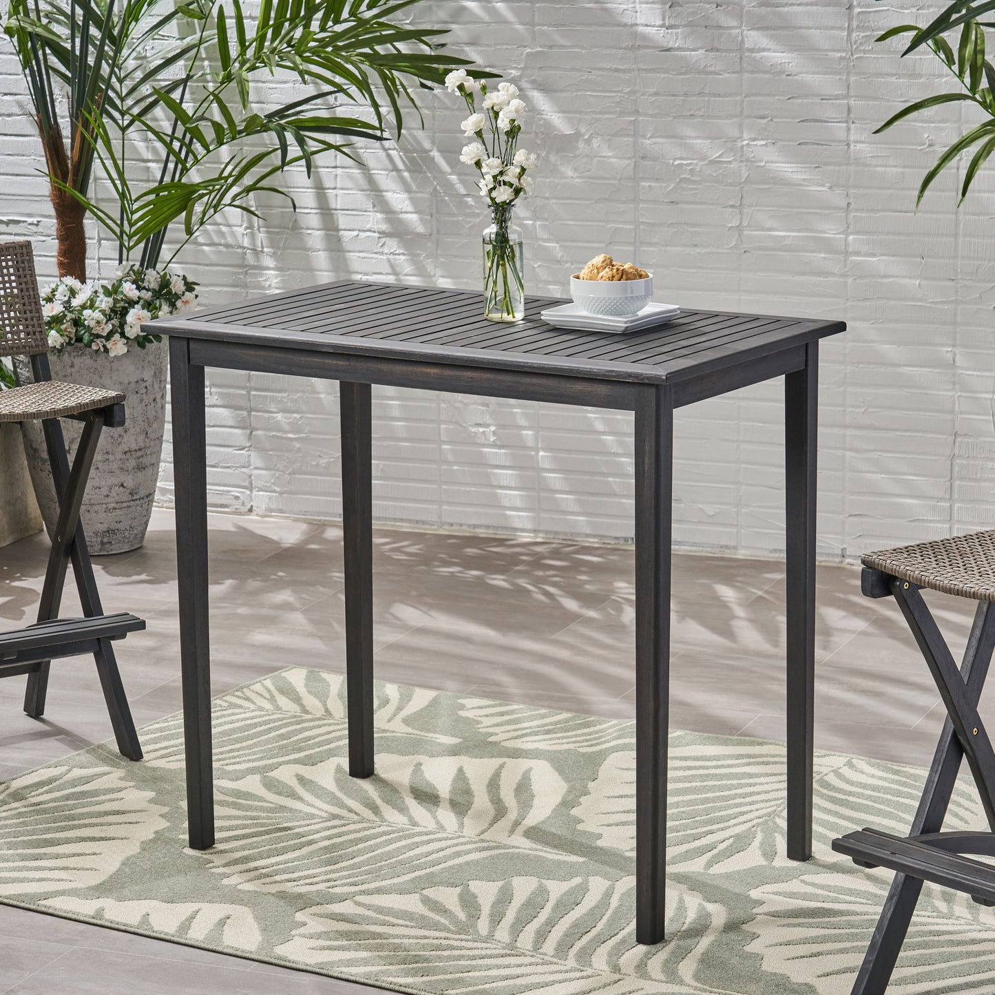 The Outdoor Minimalist Acacia Wood Rectangle Bar Table with a slatted dark wood top stands on a leaf-patterned rug, decorated with a vase of white flowers and snacks. Folding chairs and plants are arranged nearby.