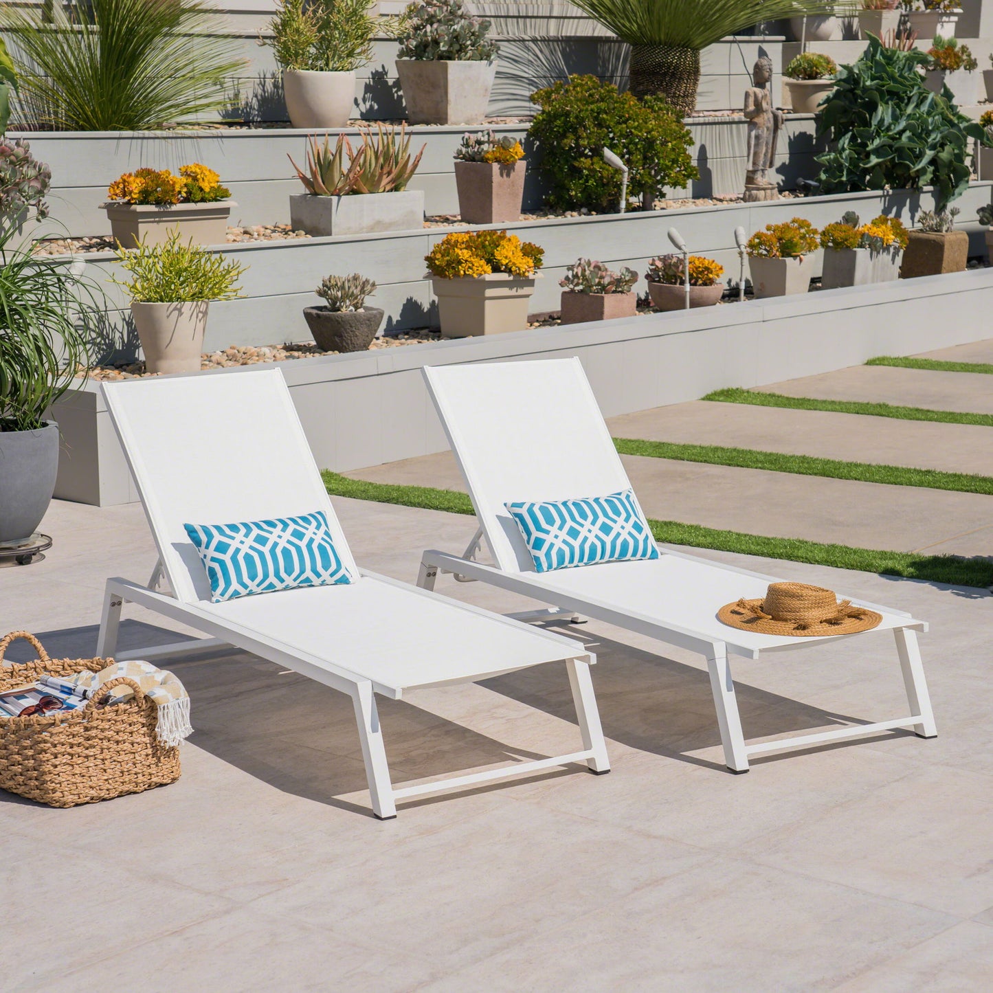 The Myers Modern Chaise Lounge Set with adjustable mesh backs, paired with blue patterned pillows and a small white table holding a straw hat, sits on a patio near potted plants, succulents on tiered shelves, and a woven basket.