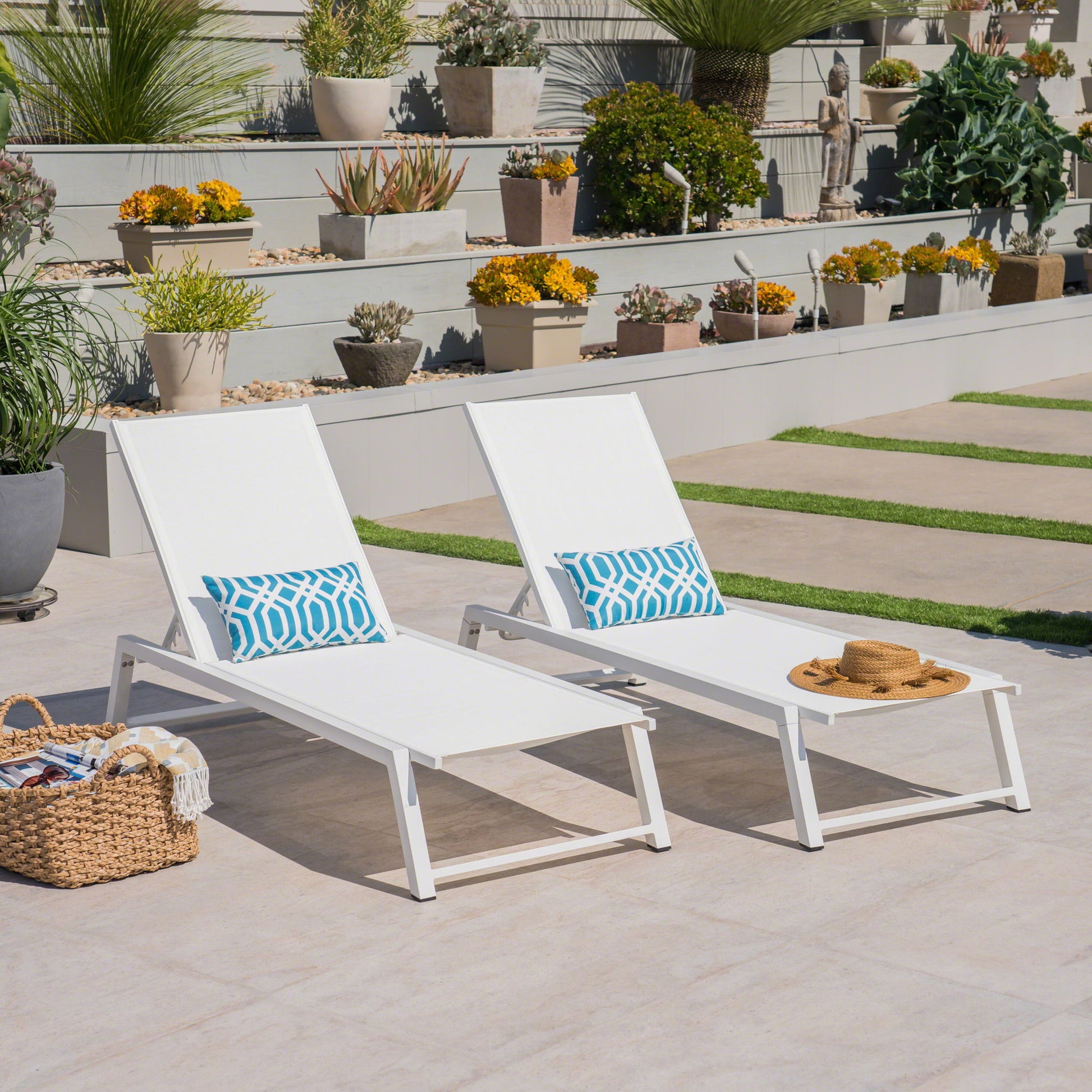 The Myers Modern Chaise Lounge Set with adjustable mesh backs, paired with blue patterned pillows and a small white table holding a straw hat, sits on a patio near potted plants, succulents on tiered shelves, and a woven basket.