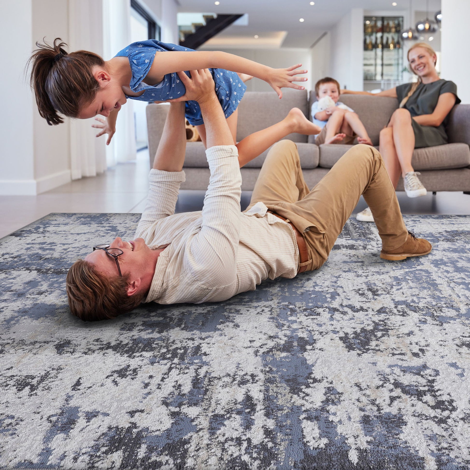 A man lifts a smiling young girl above him as they play on the Payas - Abstract Area Rug. In the background, a woman and child watch from a gray couch in a modern, well-lit living room.