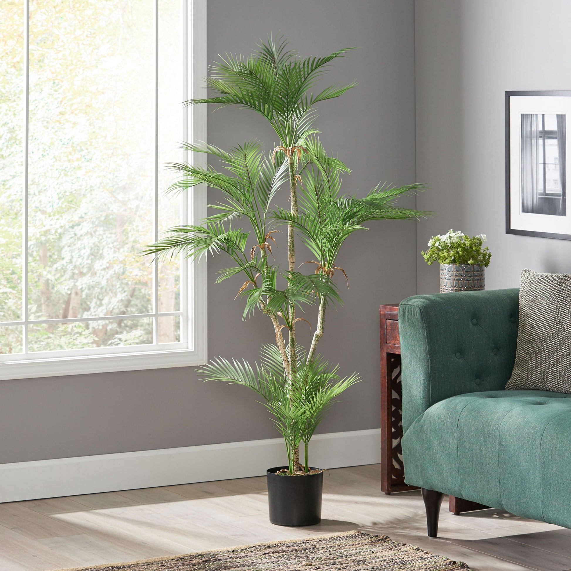 An Artificial Palm Tree is placed beside a green sofa in a bright living room with large windows, gray walls, wooden floors, and a small table with another potted plant in the background.