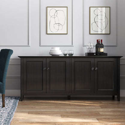 The Amherst Wide 4 Door Storage Cabinet in Hickory Brown stands against a light gray paneled wall, topped with stacked plates, glasses, wine bottles, and a decanter. Two abstract line art prints hang above it, with a blue chair partially visible nearby.