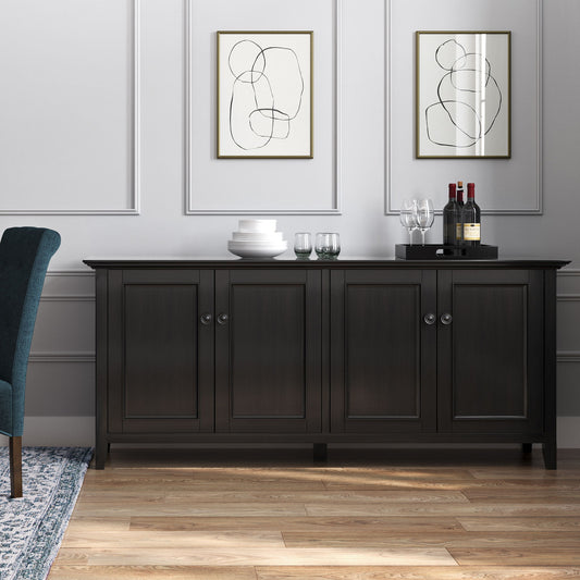 The Amherst Wide 4 Door Storage Cabinet in Hickory Brown stands against a light gray paneled wall, topped with stacked plates, glasses, wine bottles, and a decanter. Two abstract line art prints hang above it, with a blue chair partially visible nearby.