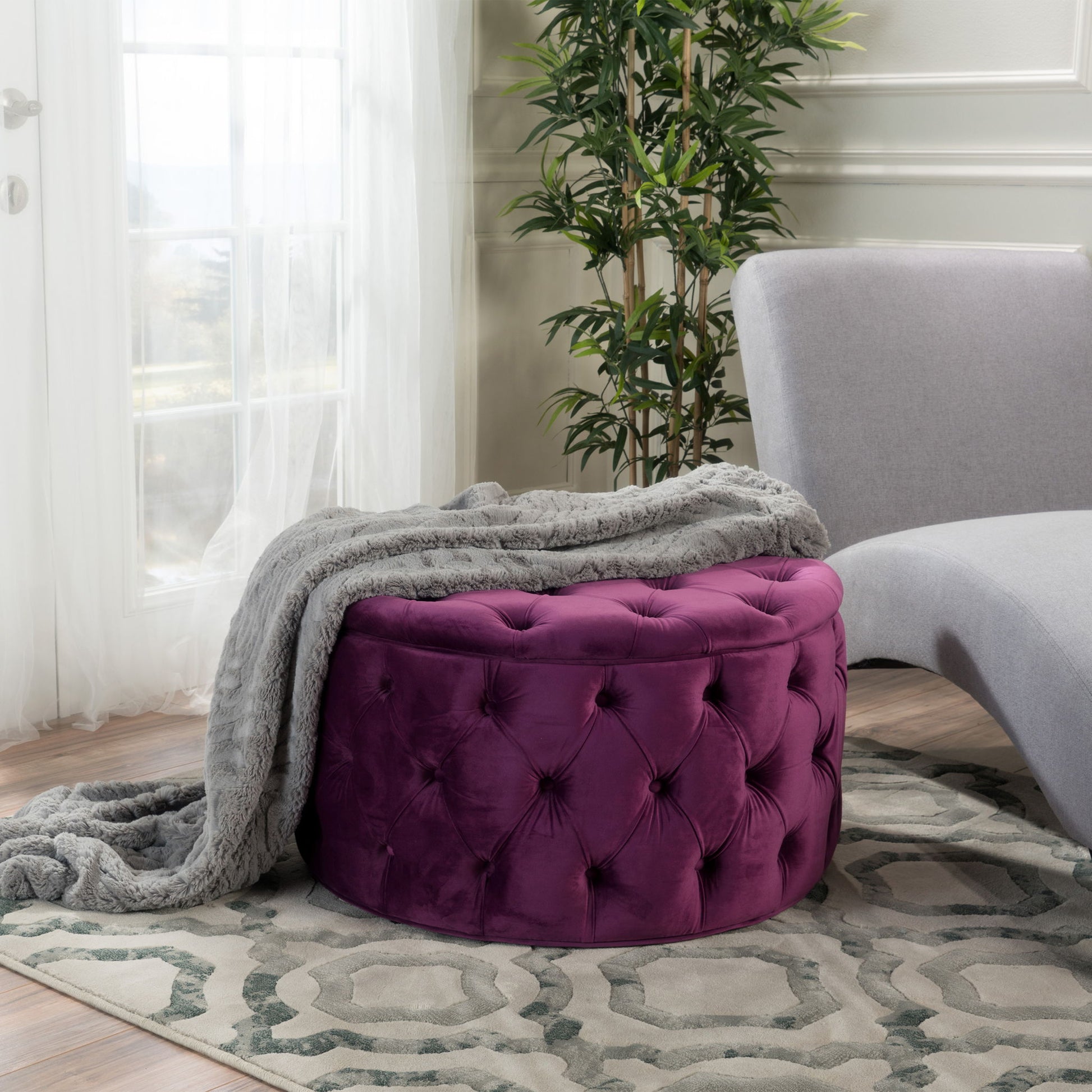 A Modern Glam Ottoman Velvet in purple sits on a patterned rug beside a modern gray chaise lounge, draped with a soft gray throw. A tall green plant and sheer curtains complete this cozy, sunlit room.