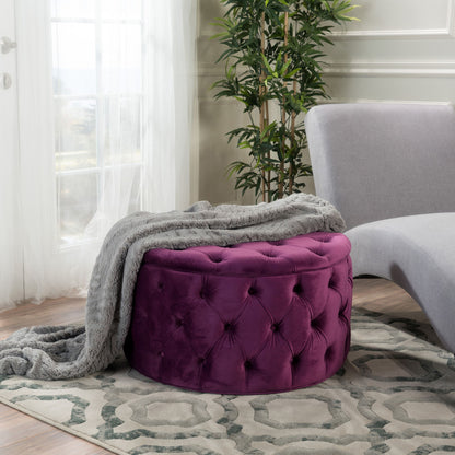 A Modern Glam Ottoman Velvet in purple sits on a patterned rug beside a modern gray chaise lounge, draped with a soft gray throw. A tall green plant and sheer curtains complete this cozy, sunlit room.