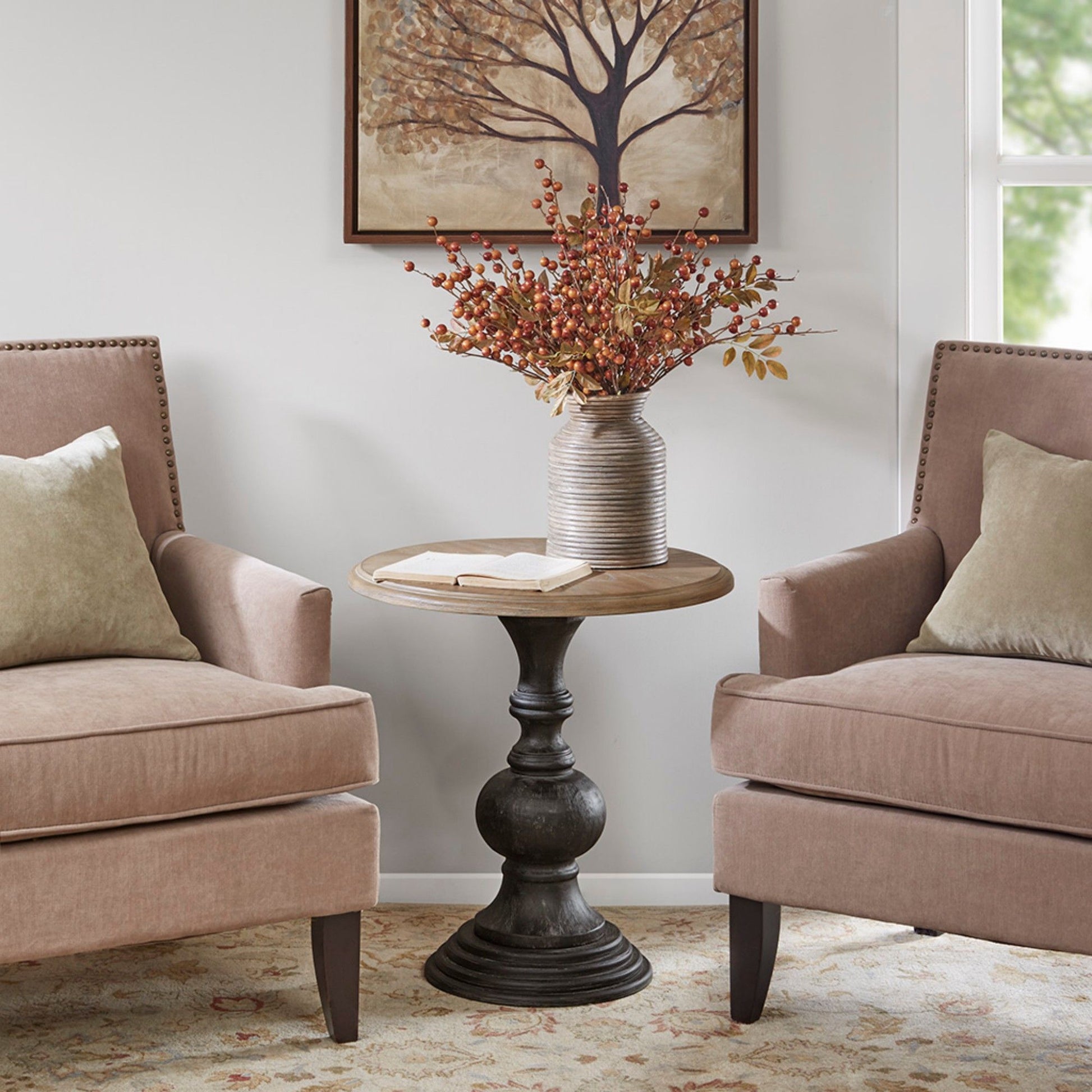 Two beige armchairs with light cushions face a Table With Reclaimed - Natural, which holds a striped vase of fall foliage and a book. A tree artwork hangs on the wall behind, illuminated by natural light.