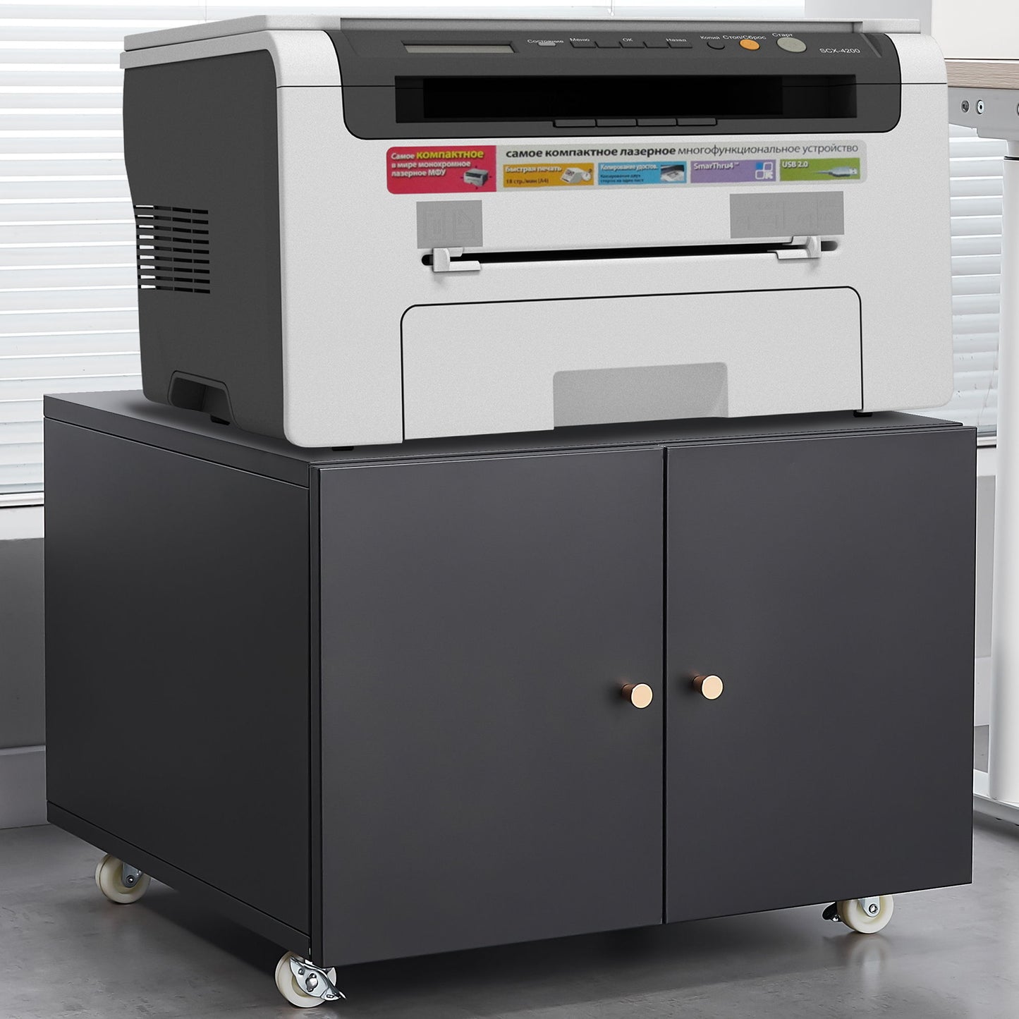 A gray and black laser printer sits on an Office Furniture Copier Cabinet 2 Door Steel Copier Stand Mobile Pedestal File Printer Stand, which features two doors and wheels, on a light gray office floor.