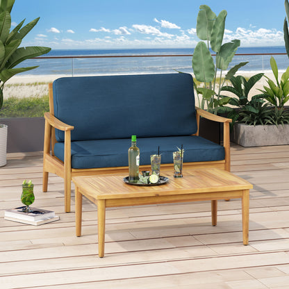 The Outdoor Acacia Wood Loveseat Set With Coffee Table, featuring blue cushions, is arranged on a deck with ocean views. The coffee table displays a tray with drinks, while lush plants surround the sunlit setting.