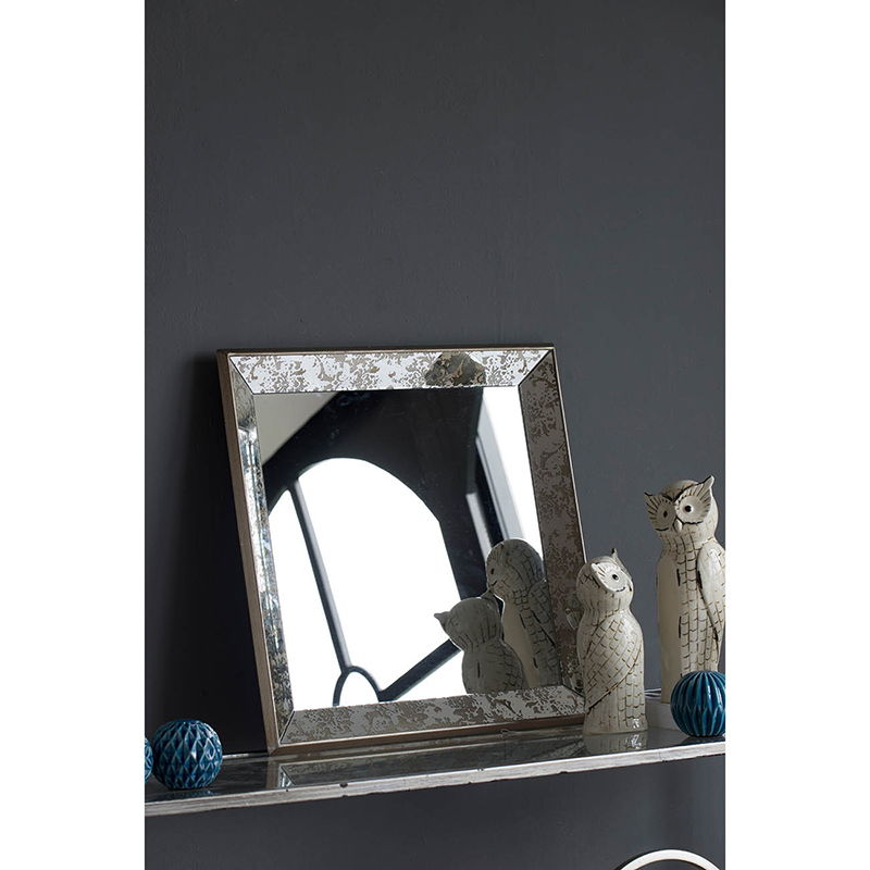 An Antique Rectangle Mirror With Floral Accents rests on a dark shelf against a charcoal wall, surrounded by ceramic owl figurines and blue textured balls, while reflecting a window with a curved frame.