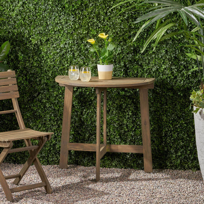 An Outdoor Half Round Folding Acacia Wood Bistro Table holds two drinks and a potted yellow flower on gravel beside a wooden chair, surrounded by green plants and a lush hedge.