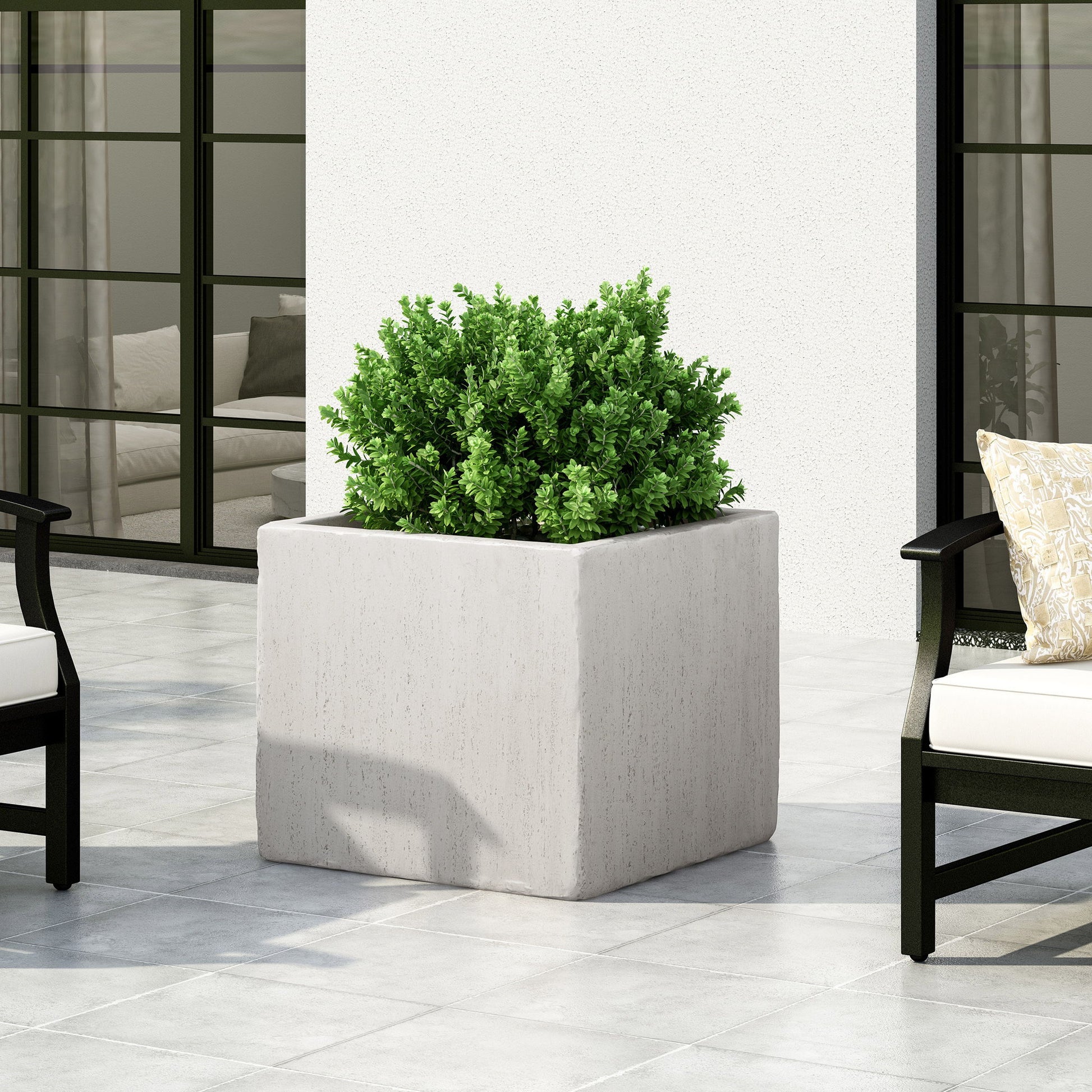 A Modern Outdoor Planter with a lush green shrub sits on a tiled patio, surrounded by sleek black chairs with white cushions, set against a white wall and expansive glass windows.