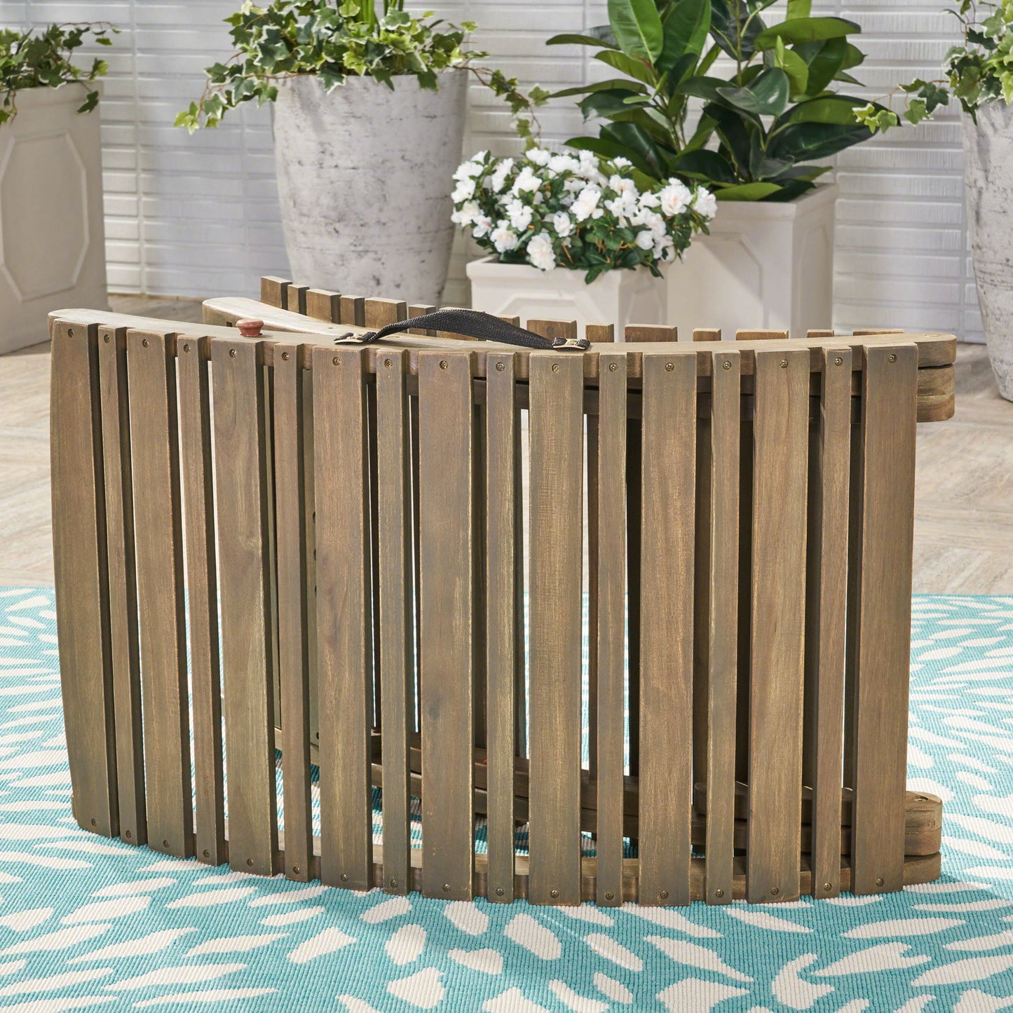 The Lahaina - Wood Foldable Chaise Lounge is displayed upright on a blue and white patterned rug, with large potted plants featuring green leaves and white flowers in the background.