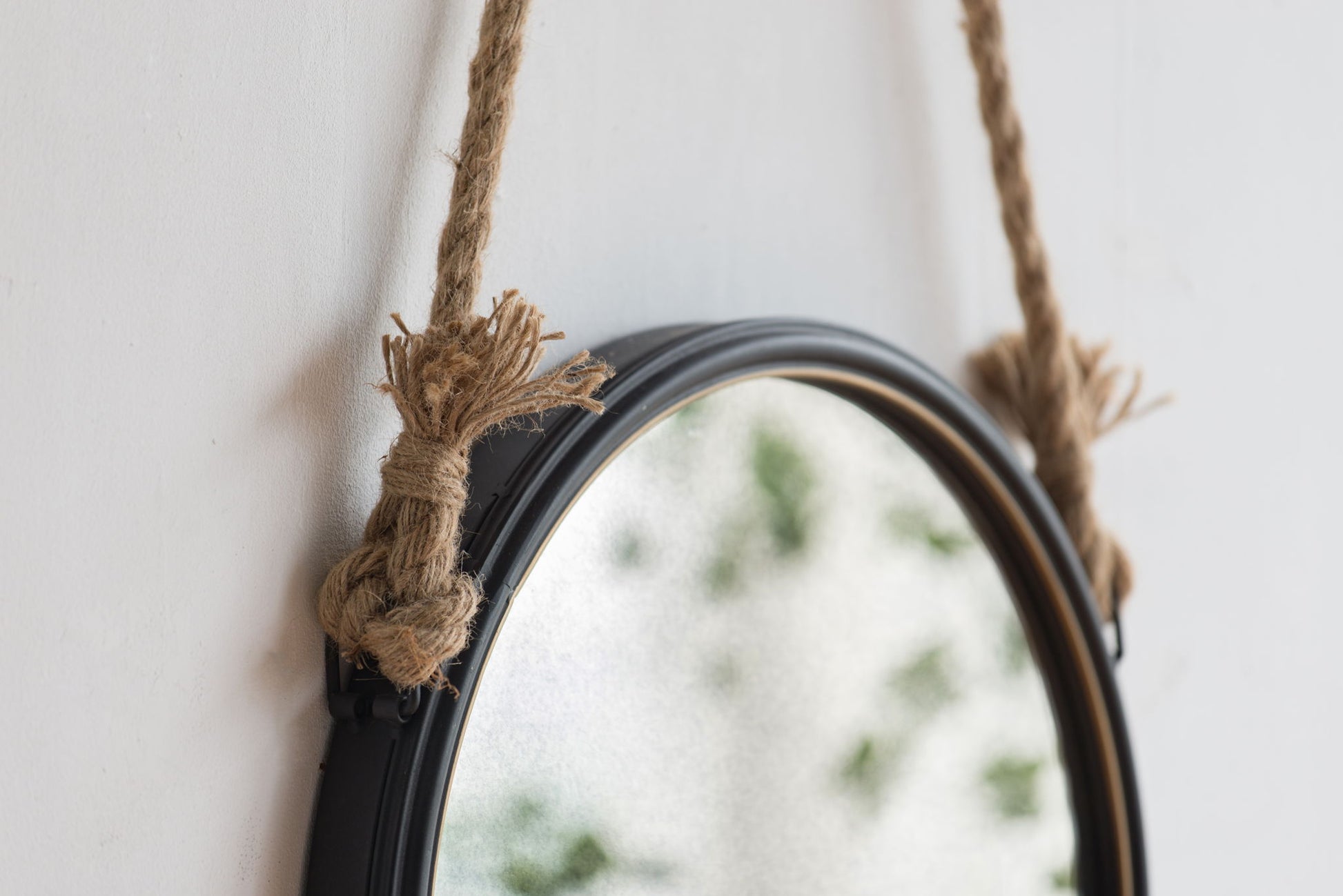 A close-up of the Handsome Cleveland Mirror With Rope Strap, a contemporary round mirror with a black metal frame, hanging on the wall and reflecting blurred greenery.