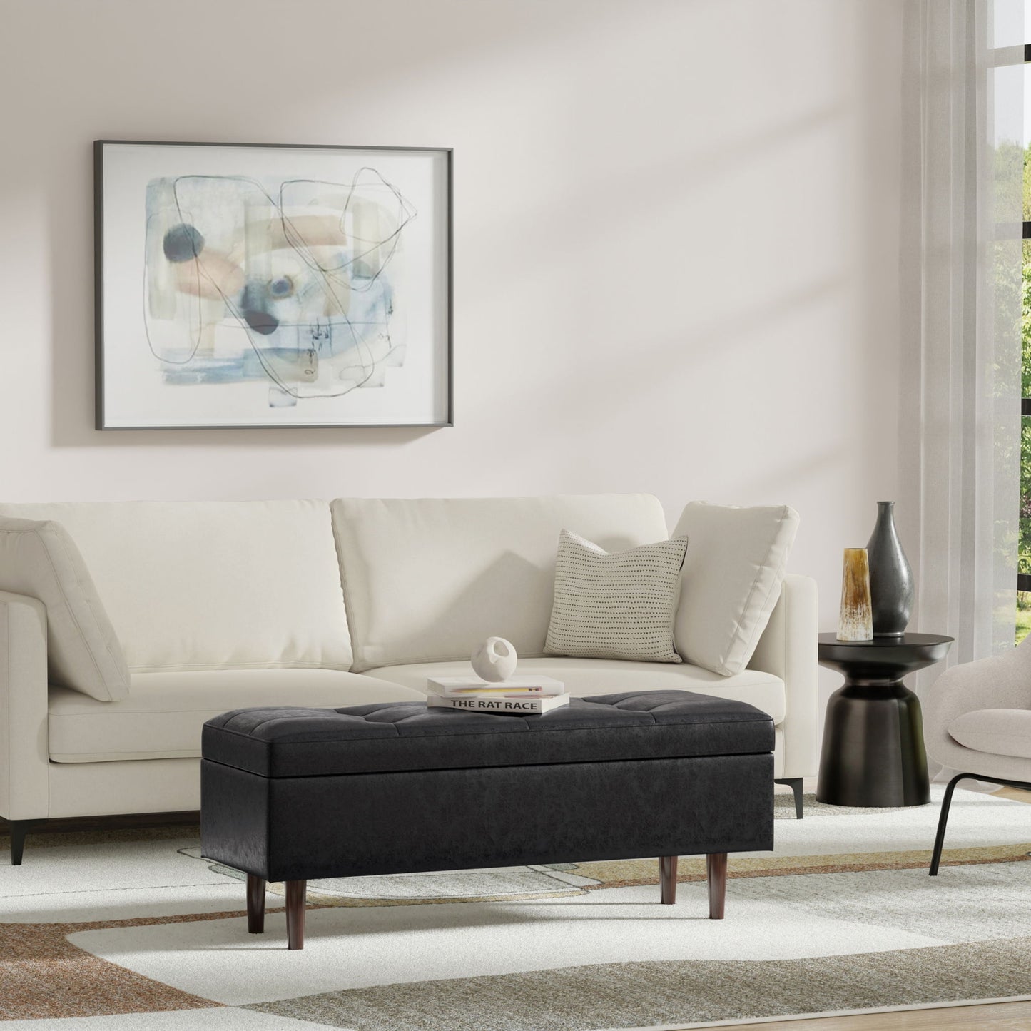 A modern living room features a white sofa, abstract wall art, the Shay Large Square Coffee Table Storage Ottoman in black, a small black side table with a vase, and a neutral rug. Sunlight pours in through the window.