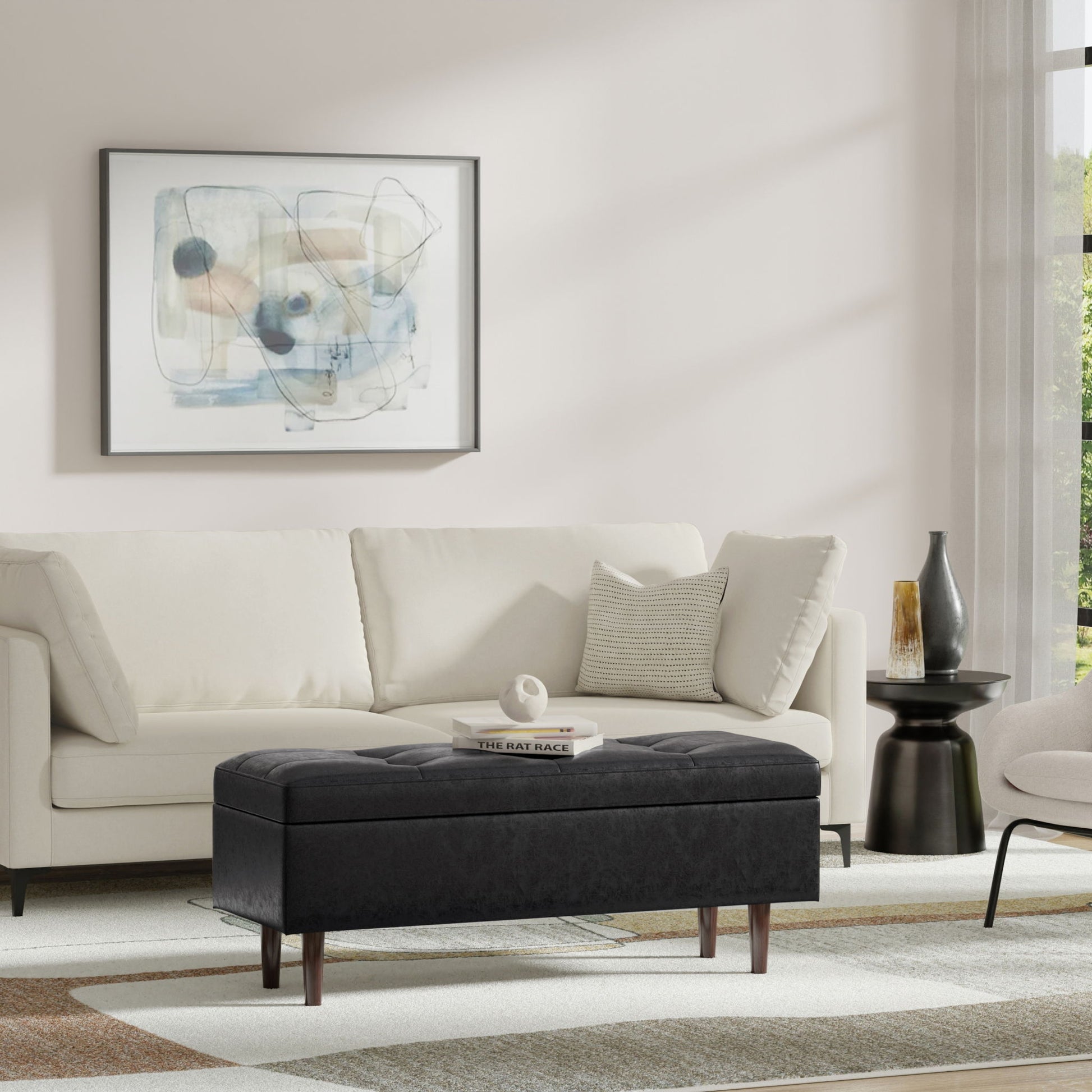 A modern living room features a white sofa, abstract wall art, the Shay Large Square Coffee Table Storage Ottoman in black, a small black side table with a vase, and a neutral rug. Sunlight pours in through the window.