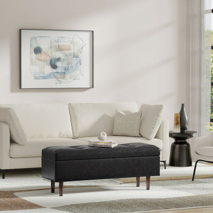 A modern living room features a white sofa, abstract wall art, the Shay Large Square Coffee Table Storage Ottoman in black, a small black side table with a vase, and a neutral rug. Sunlight pours in through the window.