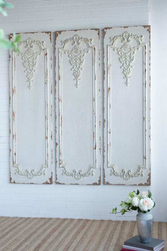 Three large wooden rectangle hanging panels with decorative carvings are displayed vertically on a white brick wall above a beige woven rug. In the corner, a vase of white roses and greenery sits atop stacked books.