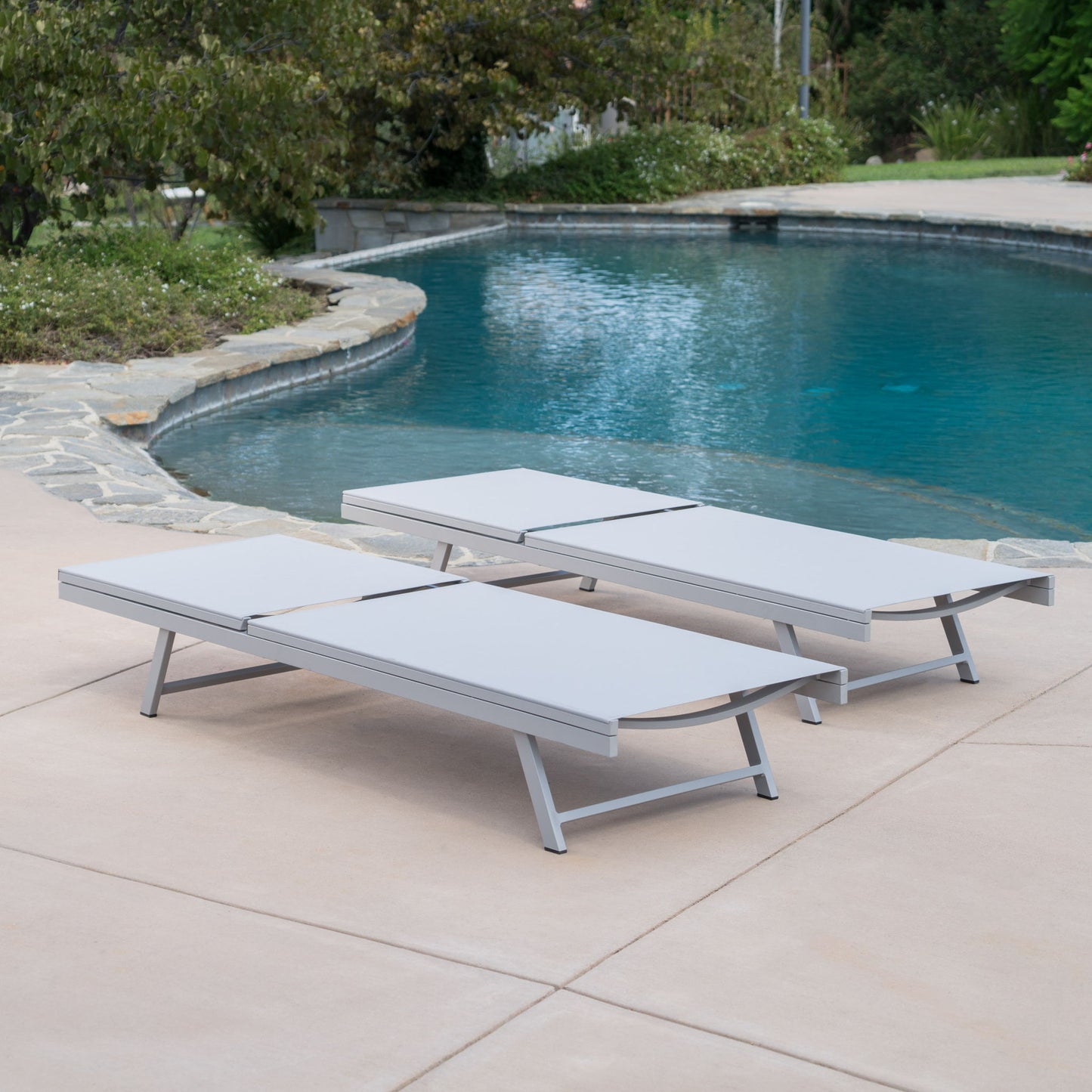 Two Salton - Chaise Lounge Outdoor Relaxation chairs sit side by side on a smooth concrete patio beside a curved pool, surrounded by trees and shrubs.