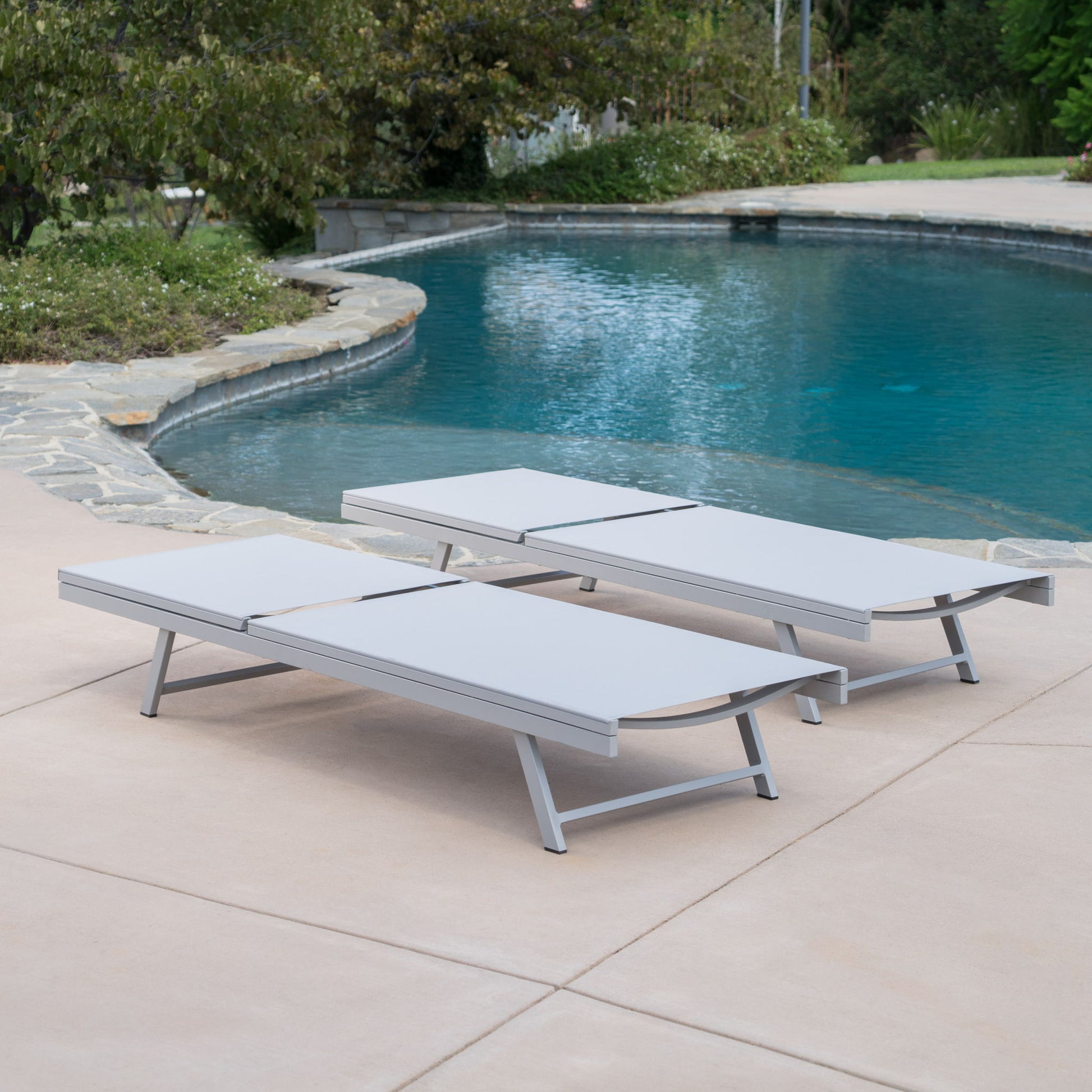 Two Salton - Chaise Lounge Outdoor Relaxation chairs sit side by side on a smooth concrete patio beside a curved pool, surrounded by trees and shrubs.