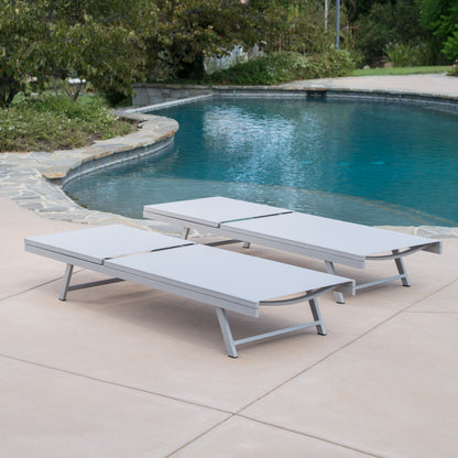 Two Salton - Chaise Lounge Outdoor Relaxation chairs sit side by side on a smooth concrete patio beside a curved pool, surrounded by trees and shrubs.